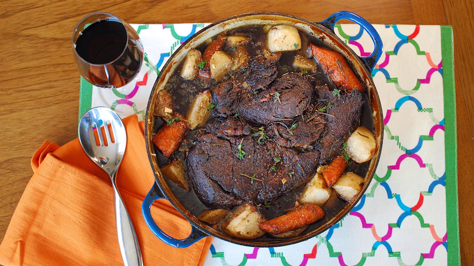 A Dutch oven containing beef pot roast with carrots, onions and turnips, accompanied by a glass of red wine.