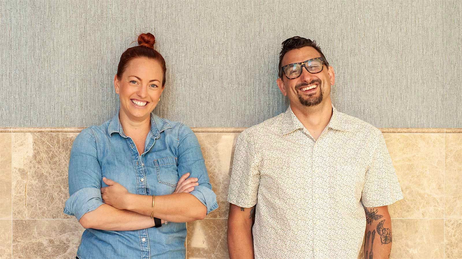The two chefs smiling at the camera, leaning against a wall