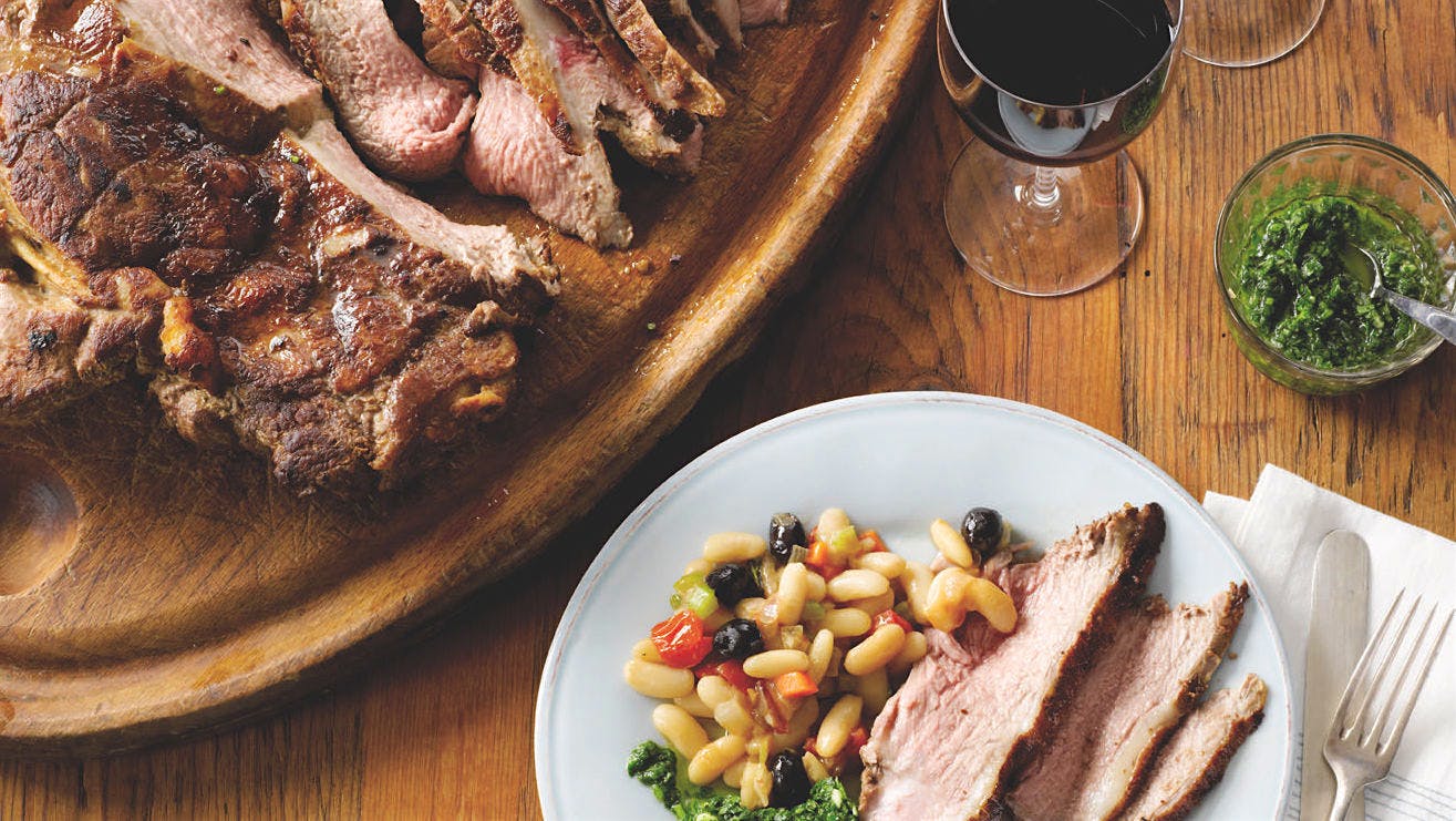 Carved leg of lamb on a platter, a bowl of white bean ragout with vegetables, a plate of lamb and beans with bright green gremolata and two glasses of red wine