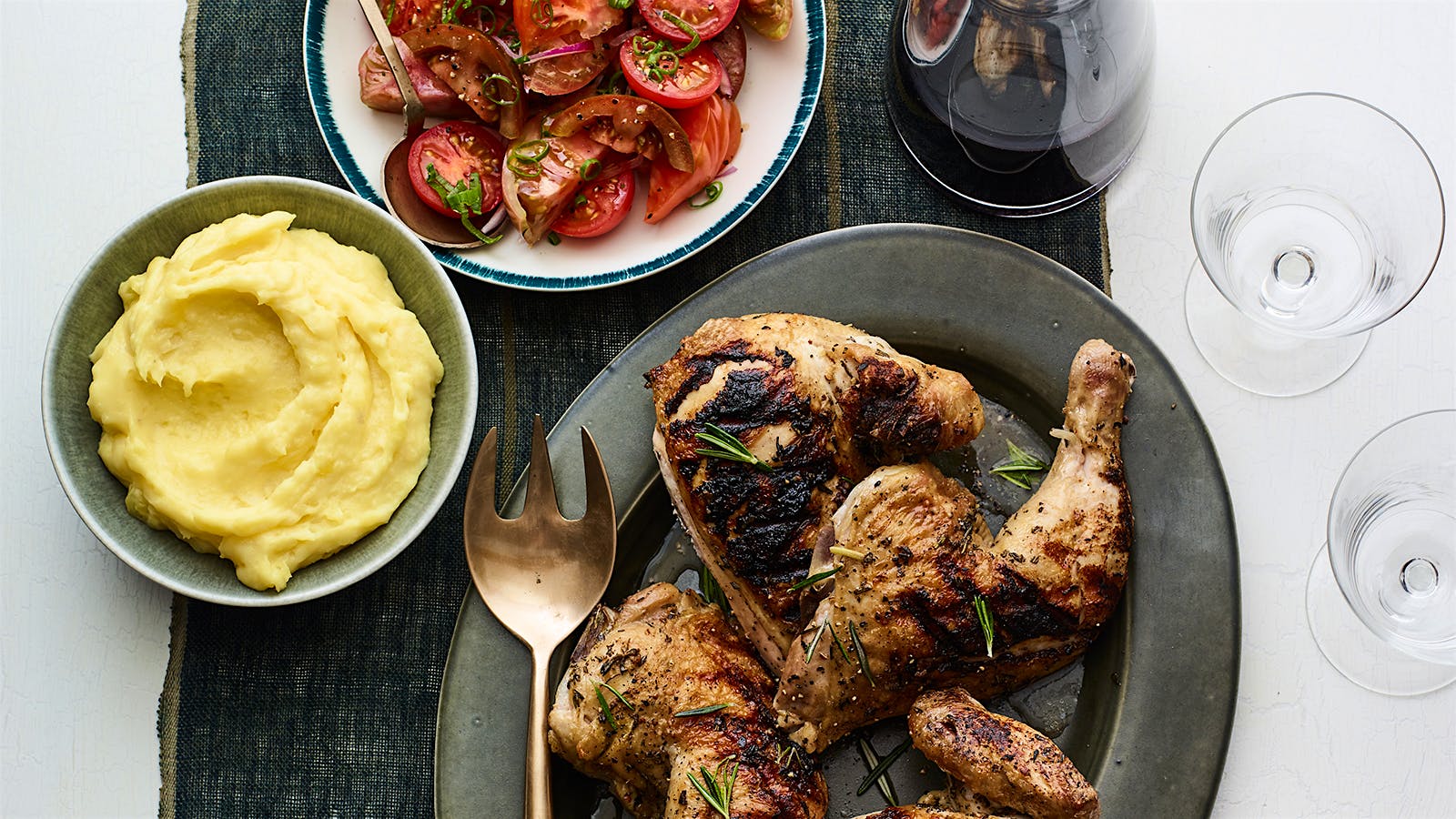 A platter of grilled chicken, a bowl of potato puree, a plate of tomato salad and a glass of red wine