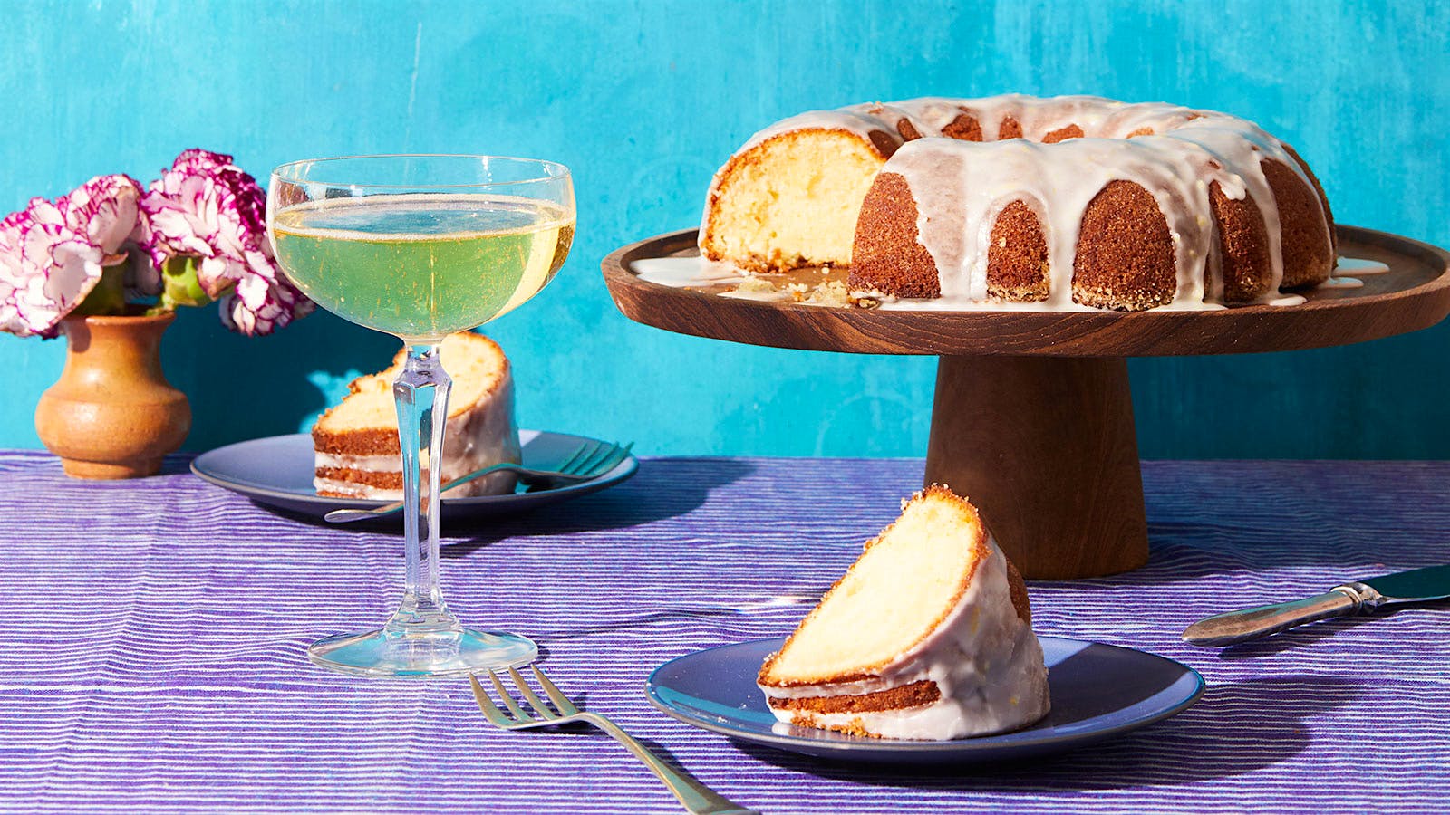 A glazed olive oil cake on a cake stand, with a coupe of sparkling wine and two slices of cake on plates
