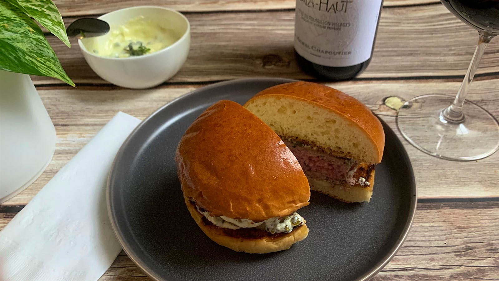 A lamb burger cut in half next to a glass and bottle of red wine and a side of yogurt sauce