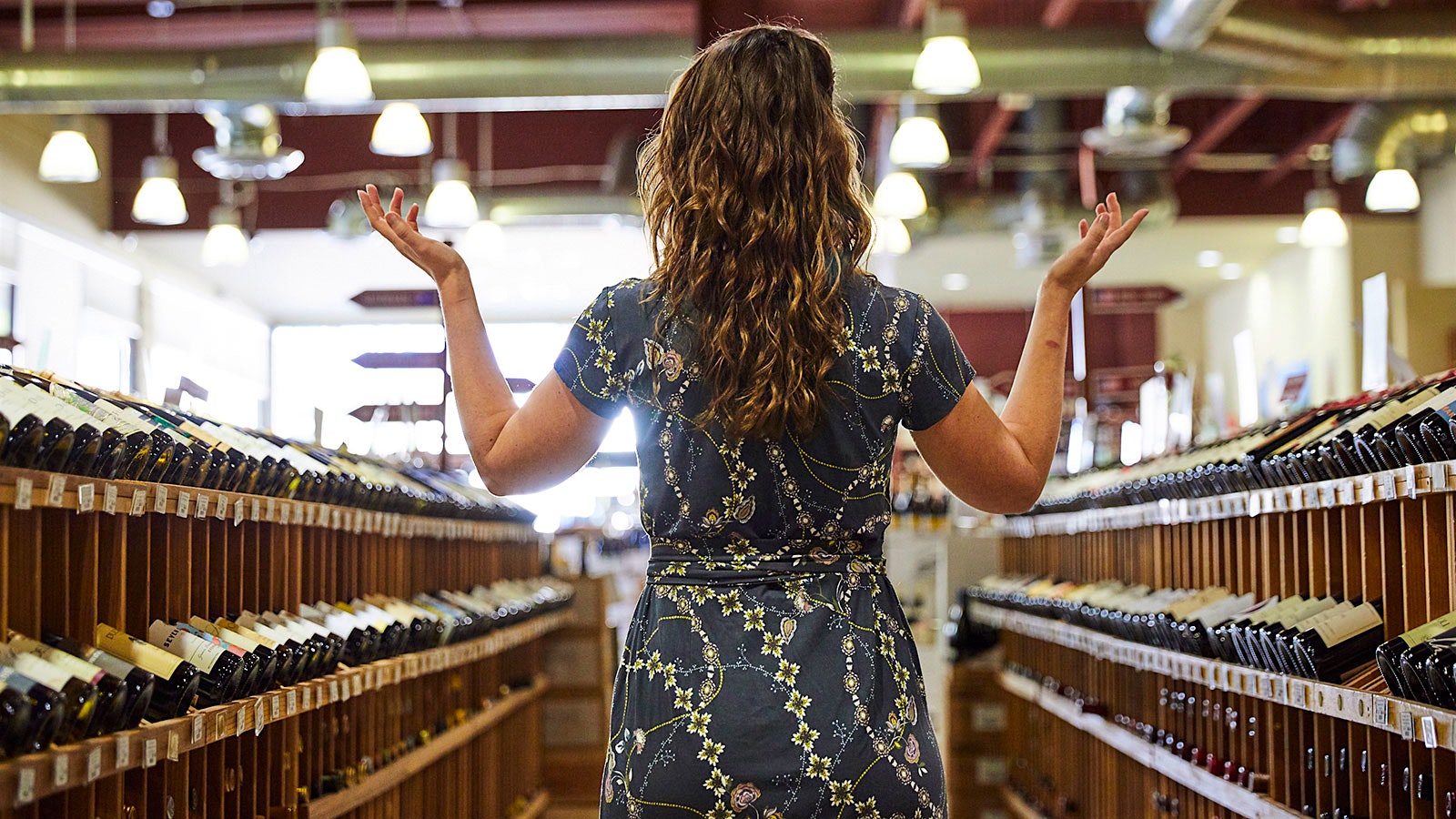 Young woman, seen from behind, throwing her hands up in the air amid the aisles of a fine wine shop