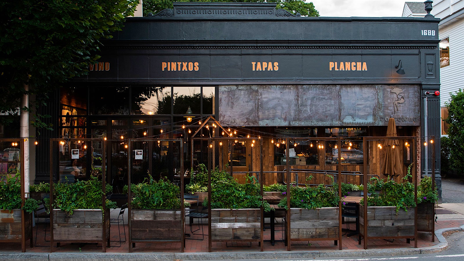  Exterior of Barcelona Wine Bar in Cambridge, Mass., with outdoor seating in front, set off by planters and decorated with string lights