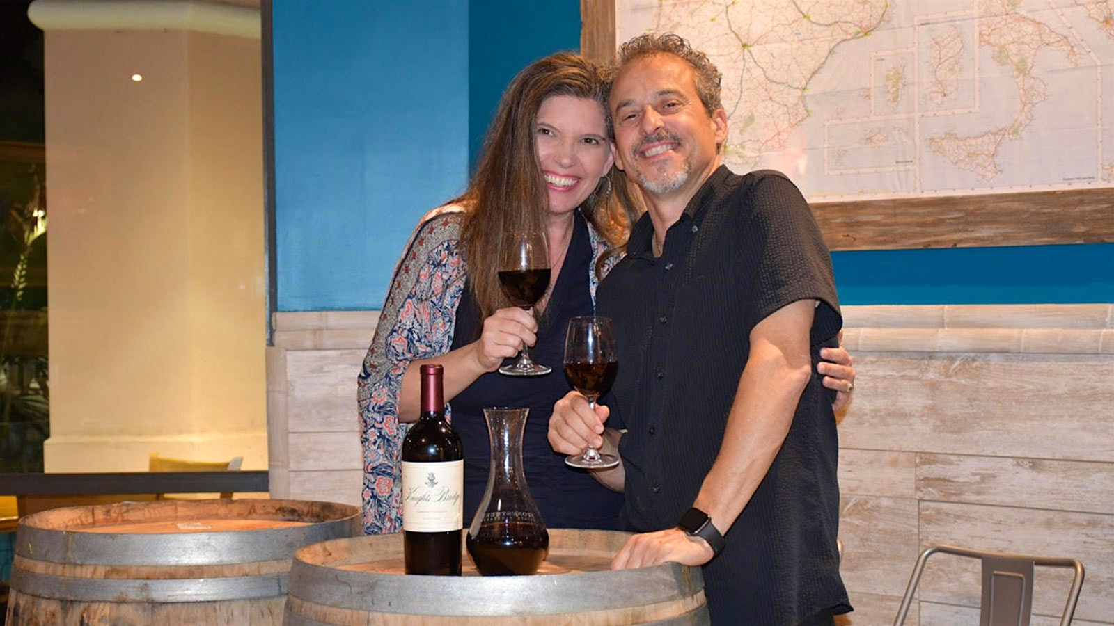   Jen et Luis Reyneri avec des verres de vin rouge derrière deux tonneaux au sommet desquels se trouvent une bouteille et une carafe de vin rouge