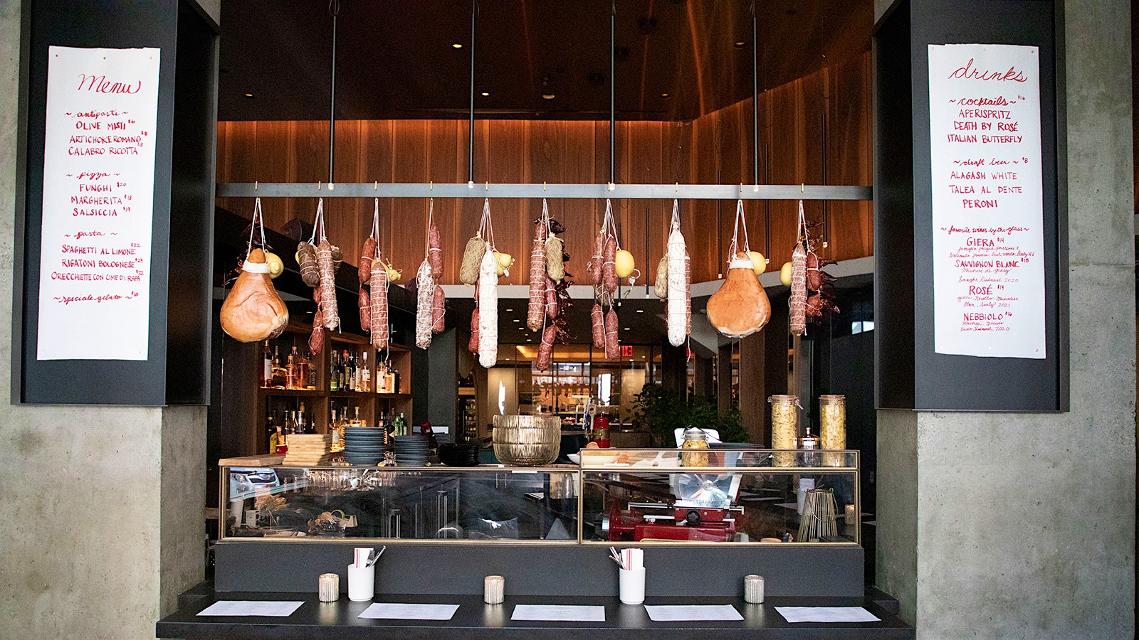  A selection of sausages, hams and other cured meats hang over a bar area with 5 white bar stools between slate-gray walls
