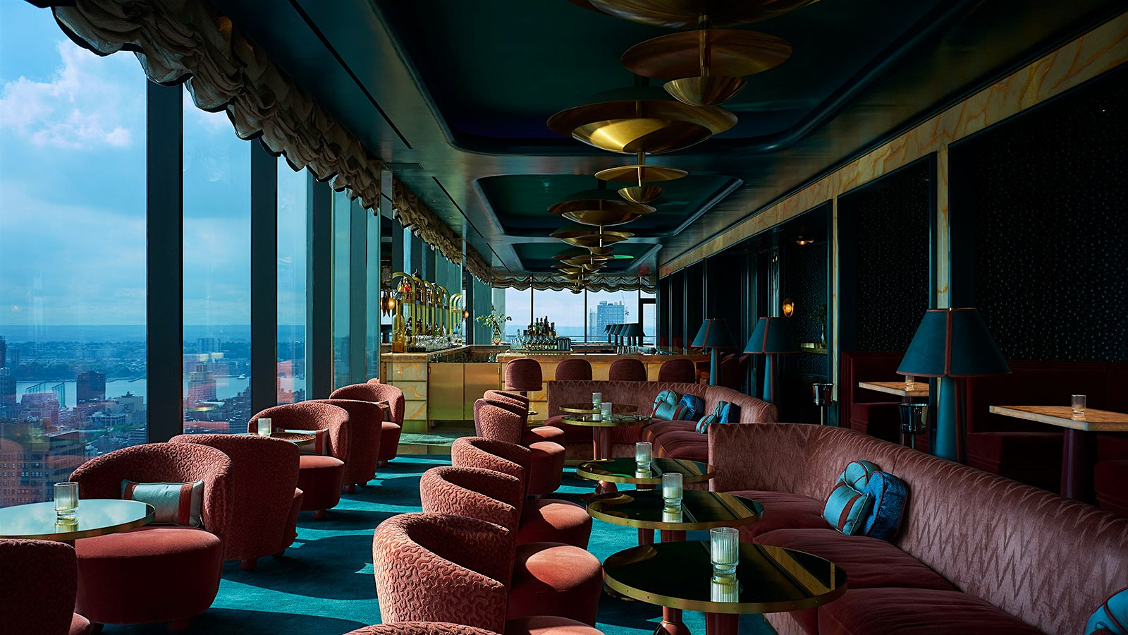  A daytime view of New York City out the floor-to-ceiling windows at Nubeluz cocktail bar, with aqua-tones, Burgundy-colored chairs and banquettes and golden light fixtures
