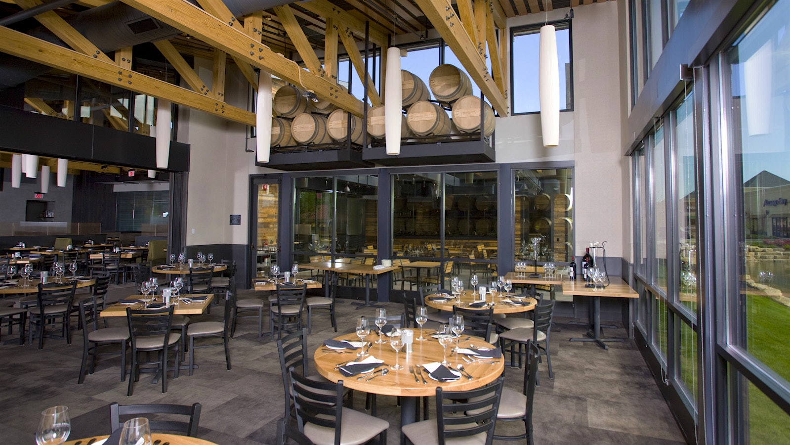  Interior of Cooper's Hawk winery and restaurant in Cline, Iowa, with dining tables, a floor-to-ceiling window, visible rafters and a glassed-in wine barrel room