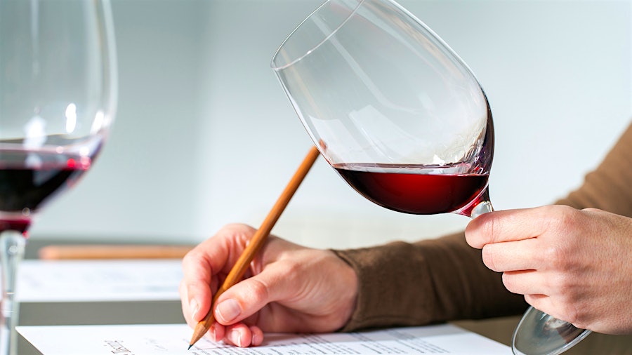 A person using a pencil to write while holding a glass of red wine; there is another glass of red wine on the person's desk.