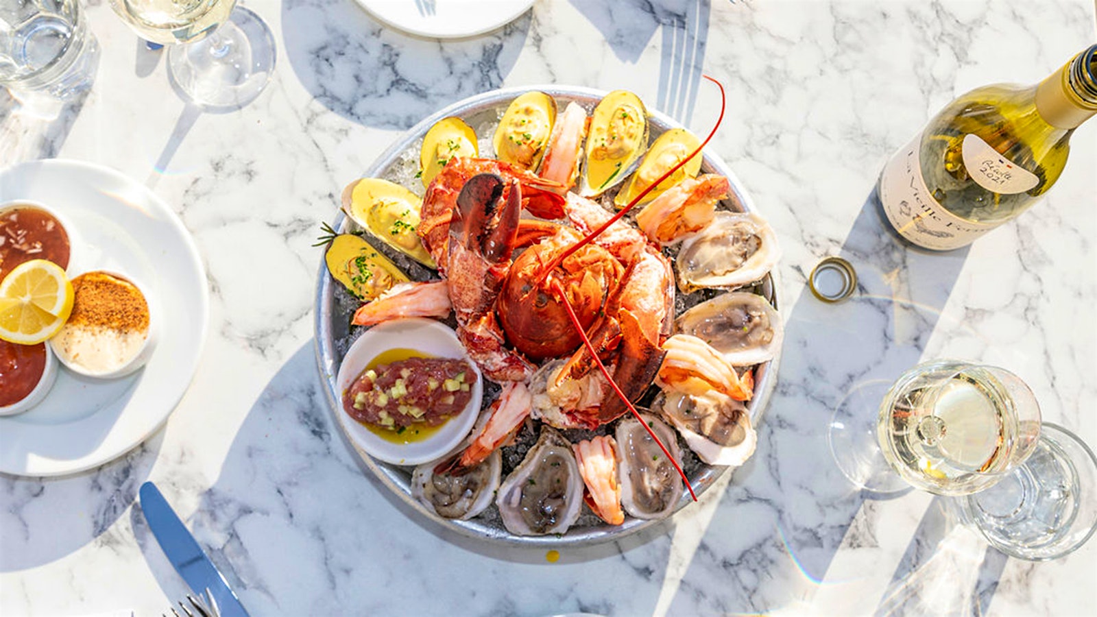  Left Bank Brasserie's shellfish platter, with lobster in the center surrounded by raw oysters, on a white marble tabletop accompanied by glasses and a bottle of white wine
