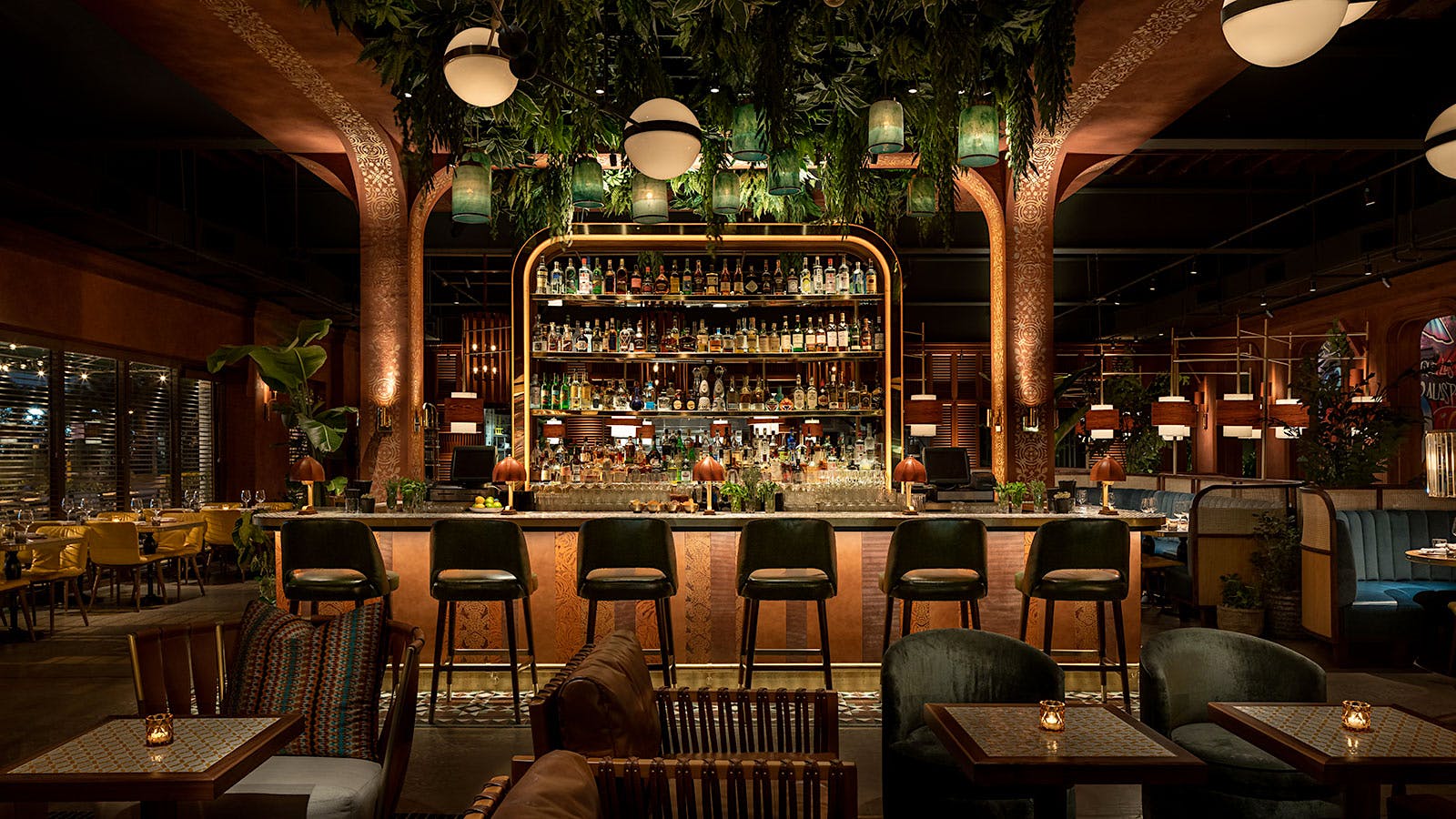  The bar and dining room at Chica, with plants hanging overhead, spheroid lighting fixtures and velvet chairs and banquets throughout