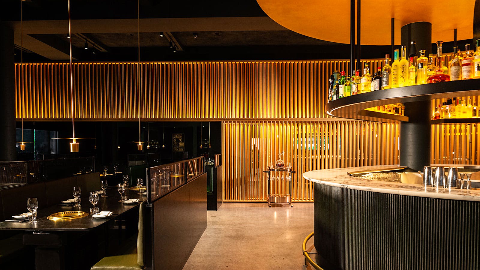  The dining room and bar at Cote Miami, with black tables and benches, grills embedded in tables, a marble bartop, bottles of spirits and a wall of wood slats