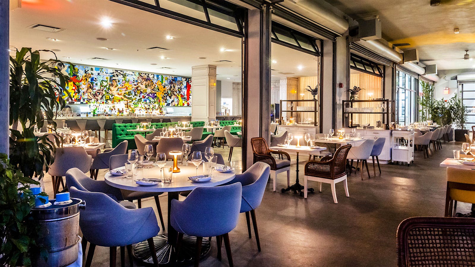  The dining room at Leku Miami, with a colorful modern art mural on the wall, blue chairs, round tables, green benches and garagelike doors separating the space