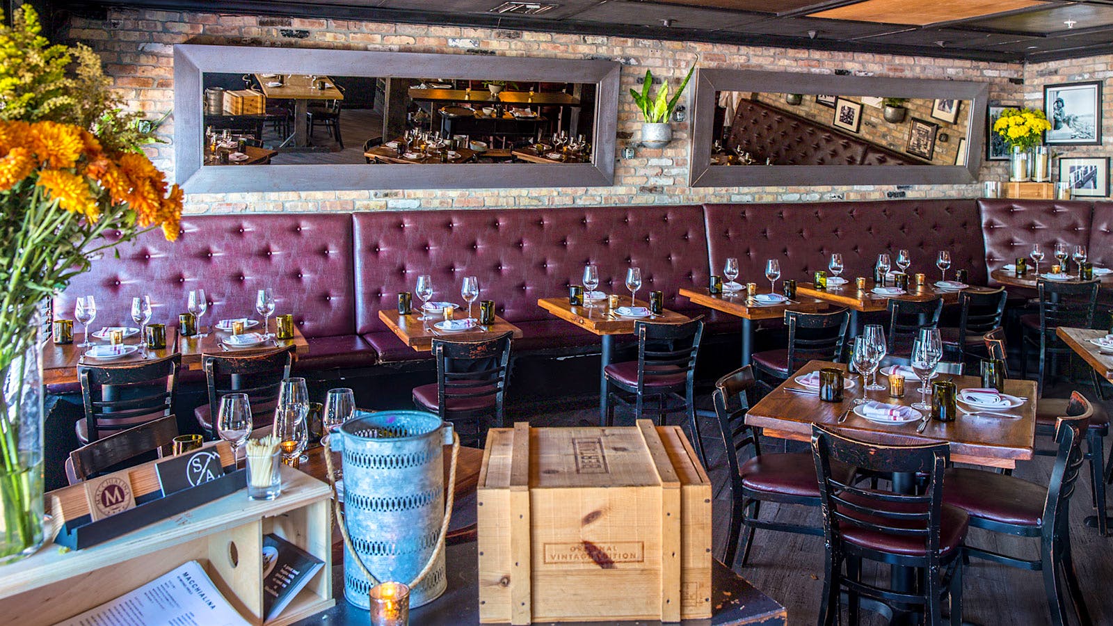  The dining room at Macchialina, with mirrors on the wall, a wood-paneled ceiling, brick wall, red leather benches and wooden crates
