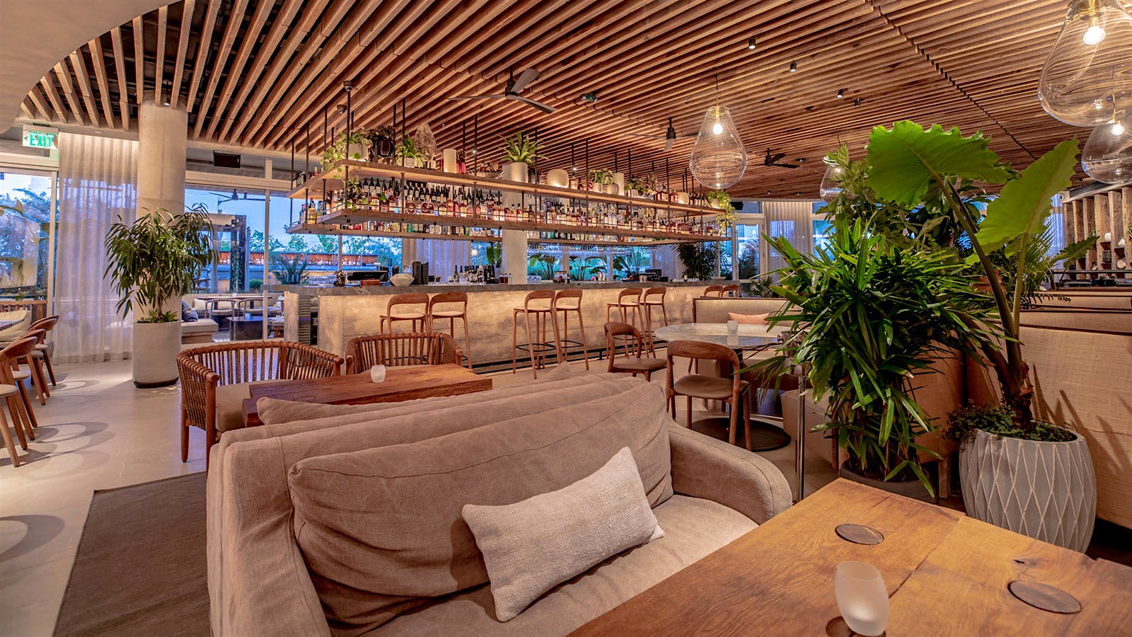  The bar and dining room at MILA Miami Beach, with stone elements, a wood-slat ceiling, wooden chairs and ceiling fans