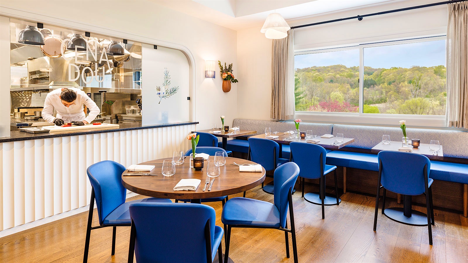  The dining room of Cenadou Bistrot, with a glassed-walled open kitchen, bright blue chairs and a view of forested hills out the window