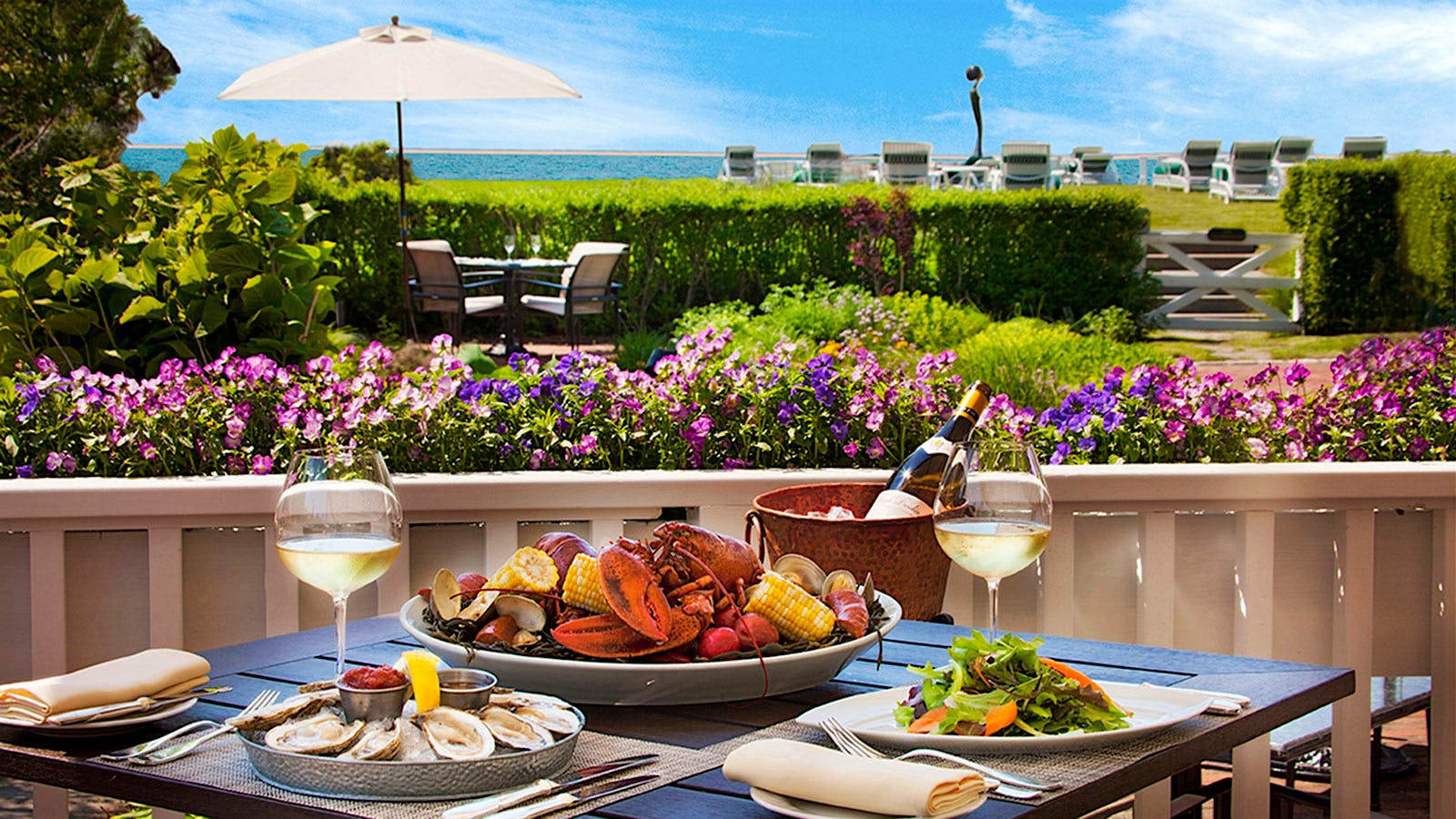 A platter of lobster, clams and corn beside a plate of salad, a platter of oysters, two glasses of white wine and a bottle of white wine in an ice bucket on a set table outside at Topper's at the Wauwinet