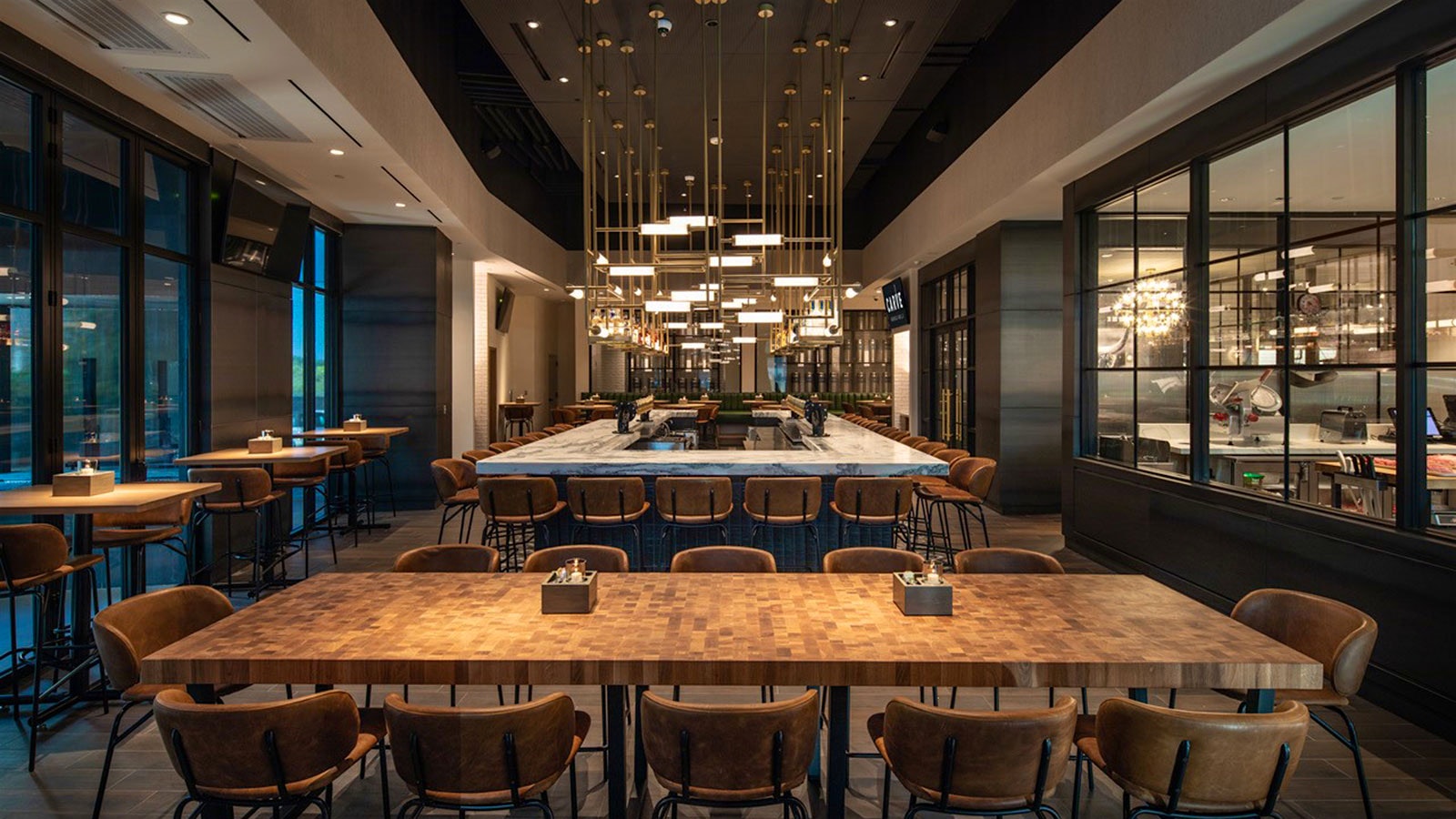  The bar area at Carve American Grille in central Austin with a rectangular marble bar with dangling lights overhead, two-person bar tables along the windows, a large 12-person table in front, and a glassed-in kitchen area to the right