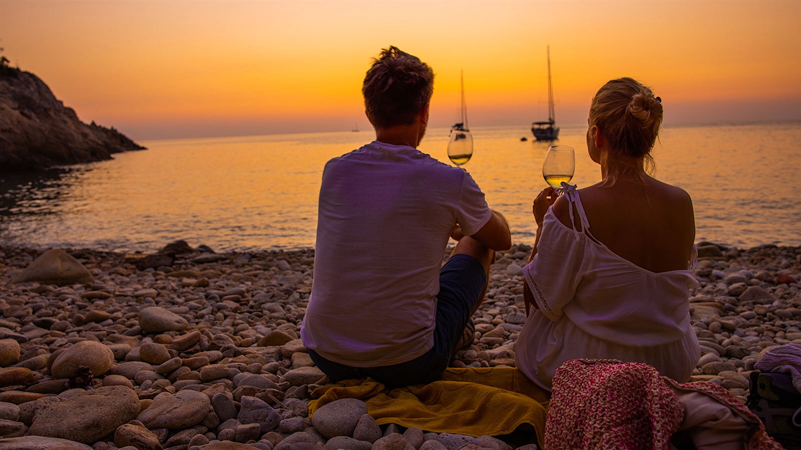  A couple sitting at the beach with glasses of white wine, looking at the sunset