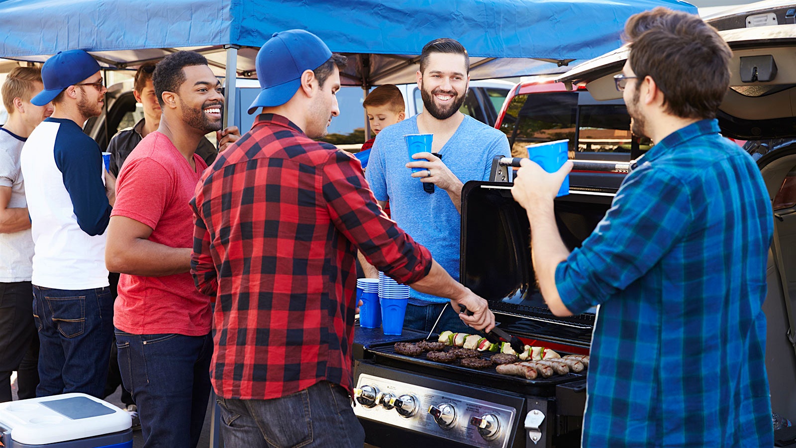  Group of men at a tailgate gathered around a grill, drinking out of plastic cups