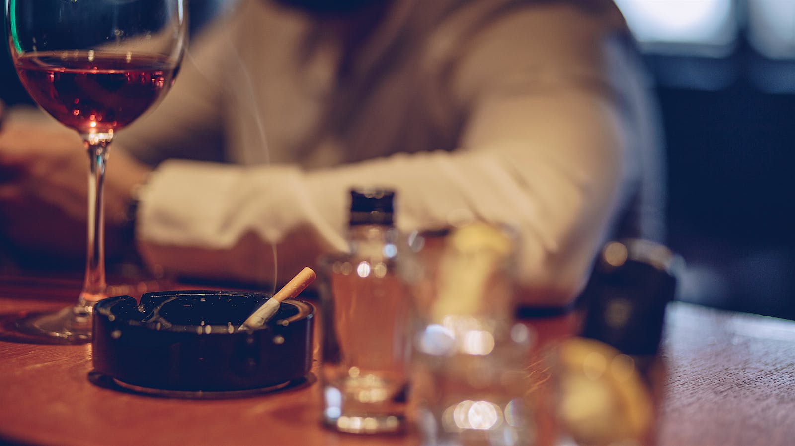 A man sitting at a bar with a glass of red wine and a lit cigarette in an ashtray