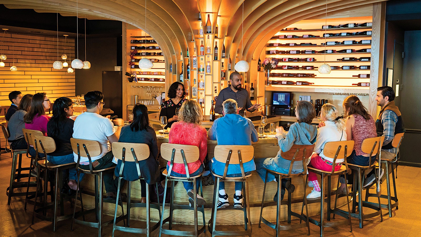  Swati Bose and Kabir Amir leading a tasting for guests at Flight Wine Bar at a wood bar with wood chairs in a room with other wood elements and wine bottles on wood shelves