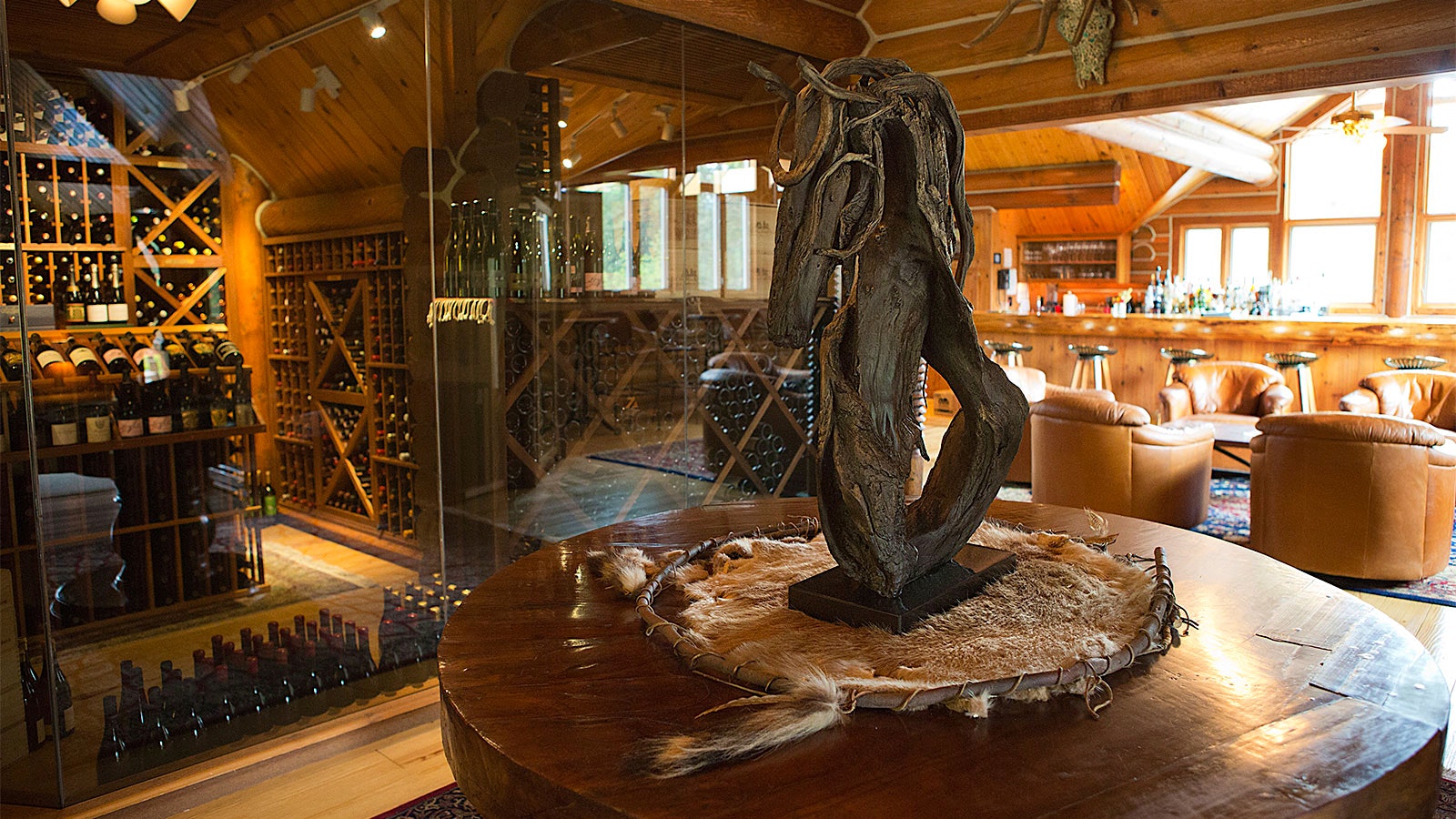  A glass-encased cellar of wine bottles at Triple Creek Ranch near leather seats, a bar and a rustic wood sculpture on a table