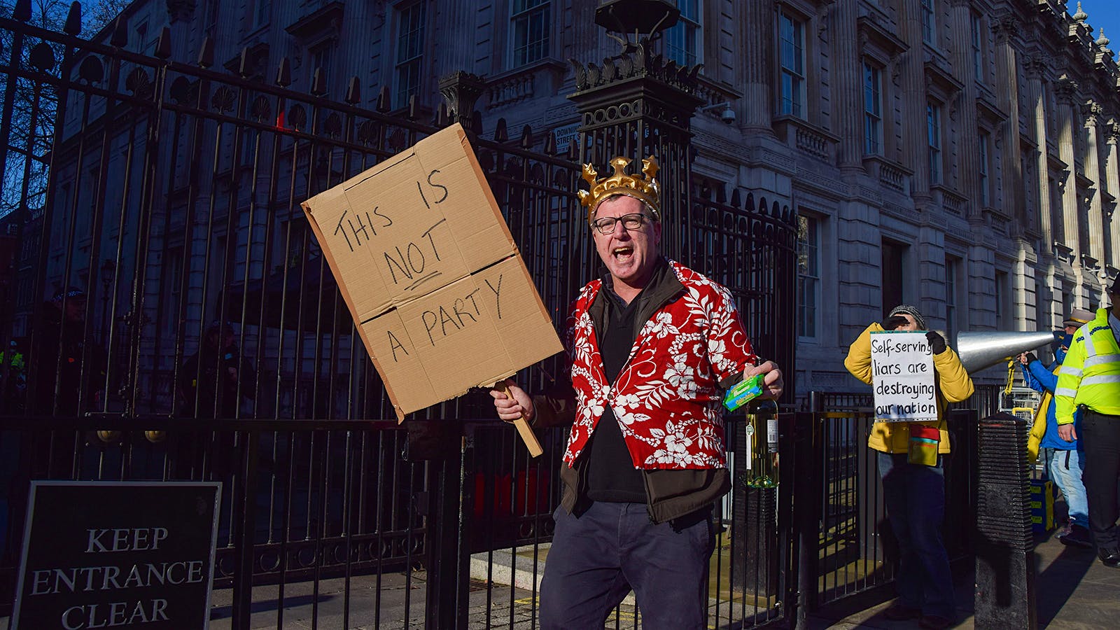 A protester wearing a crown holds a packet of cheese, a bottle of wine and a 'This Is Not A Party' placard chants during the demonstration outside Downing Street.