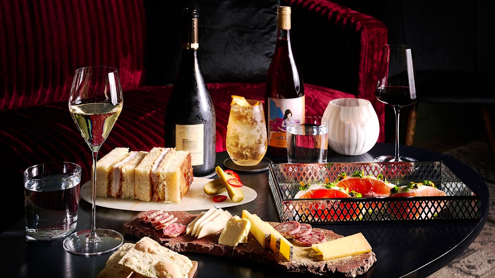  A table with red and white wine, displayed in bottles and poured in two glasses, alongside cocktails, finger sandwhiches and charcuterie boards.