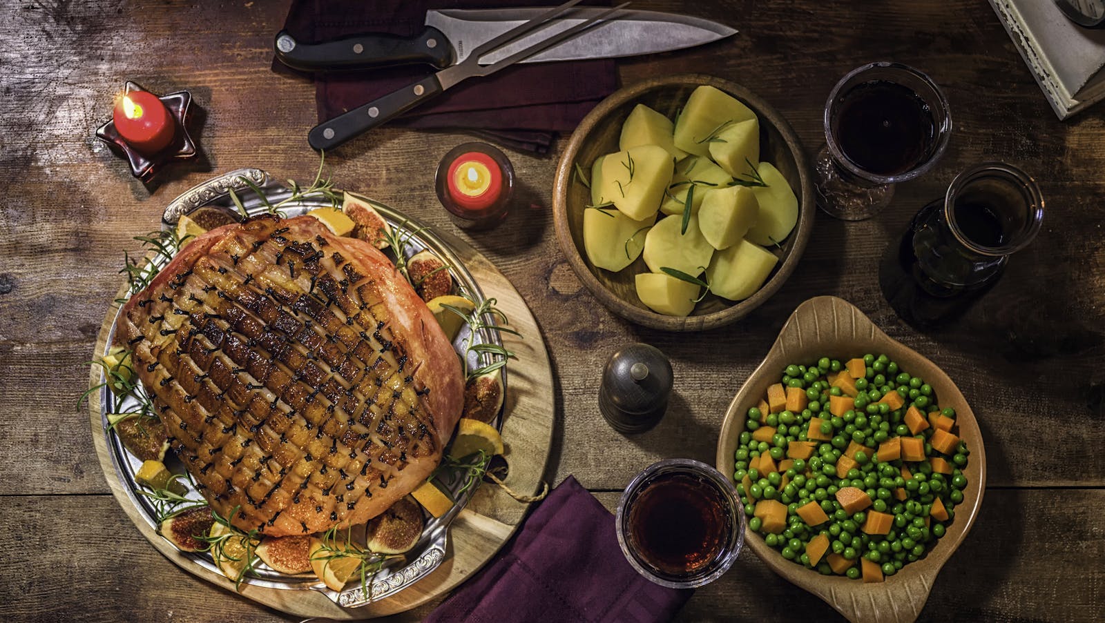 A plate of clove-studded glazed ham accompanied by side dishes, glasses of red wine and carving knives on a wood table