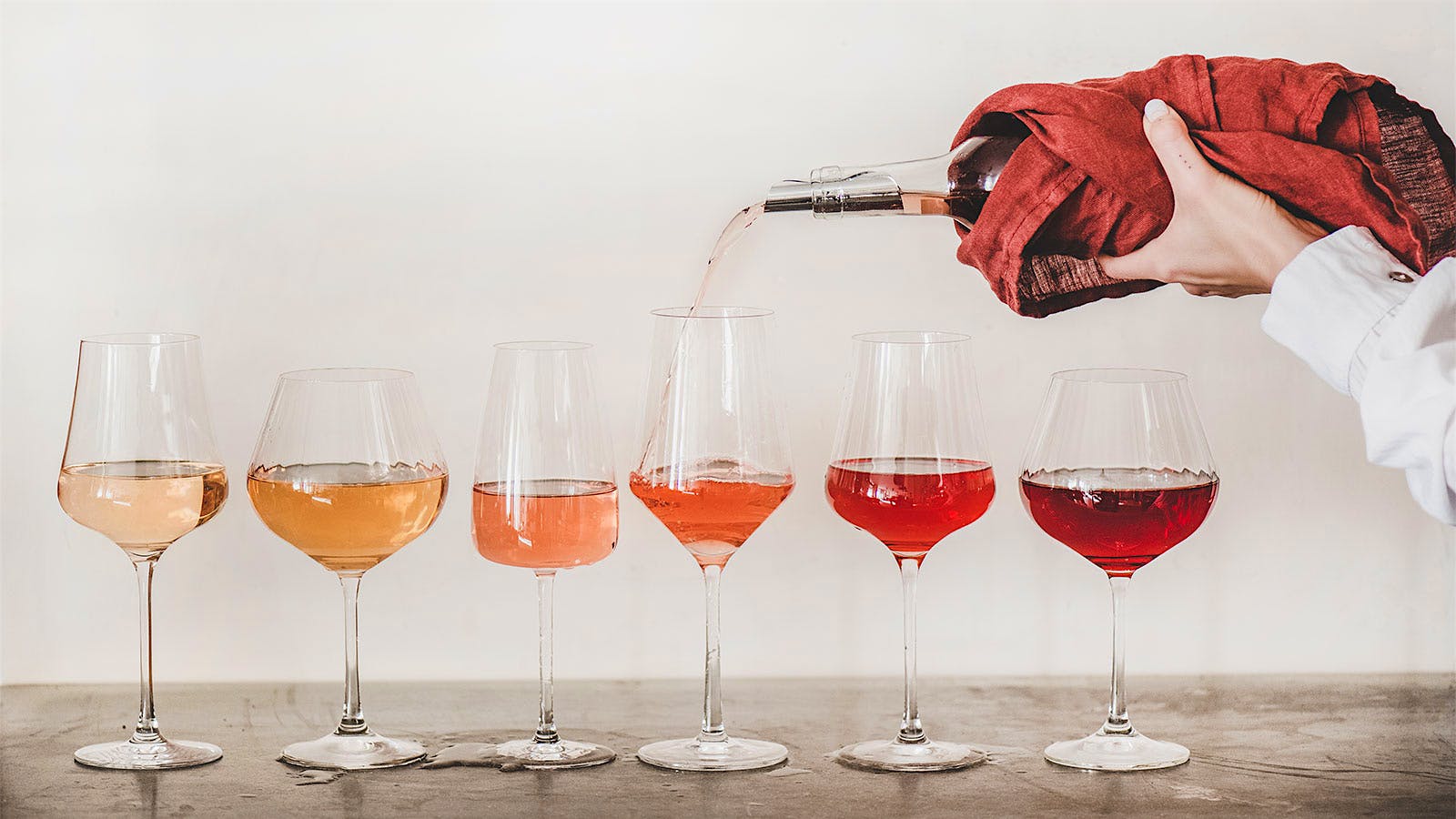 A lineup of glasses of rosé in different shades, from pale pink to light red, with a bottle being poured into one glass