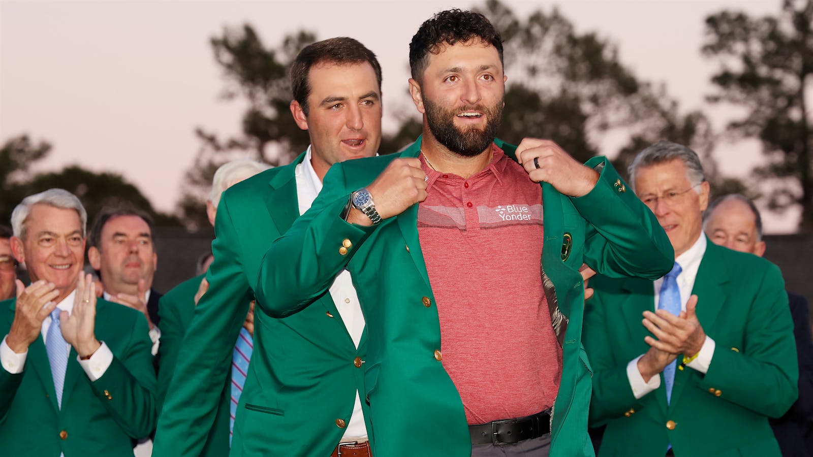 Golfer Scottie Scheffler helps put the Champion's green jacket on Jon Rahm at the 2023 Masters Tournament, as other former Champions applaud.