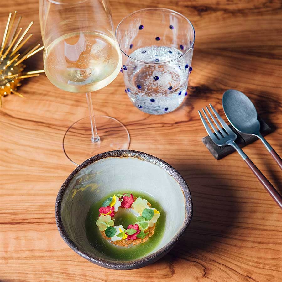  A light wood table at The Yeatman holding a glass of white wine, a clear water tumbler with blue dots and a ceramic bowl of artistically plated Galician scallops with green apple