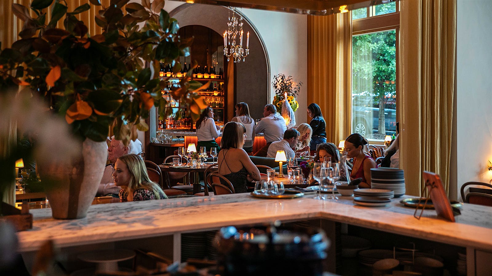 People enjoying wine and food at Le Bon Nosh, where there are white walls and yellow curtains in front of a large window