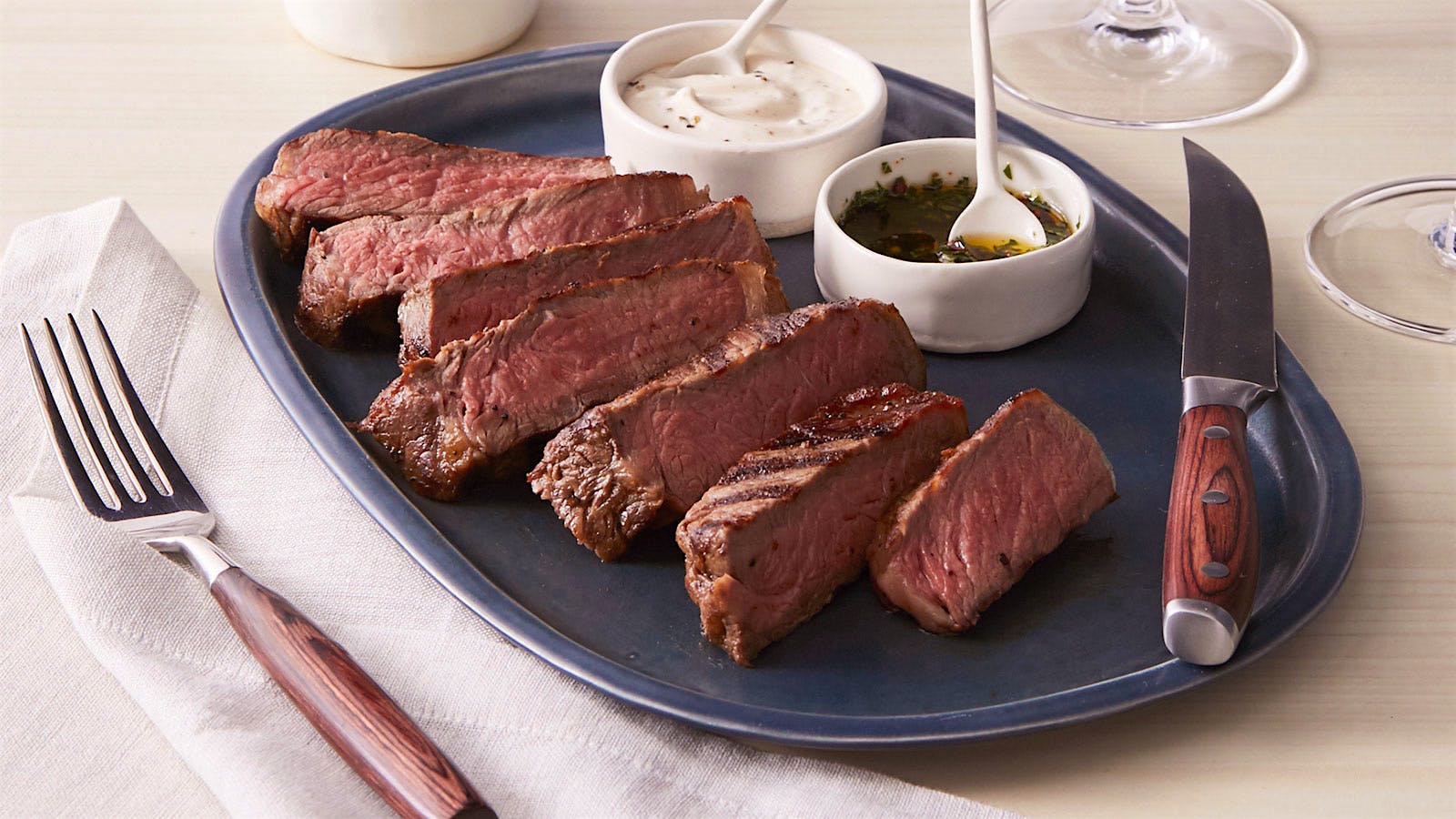 Sliced steak on a platter with a wood-handled knife, sauce ramekins and wine glasses