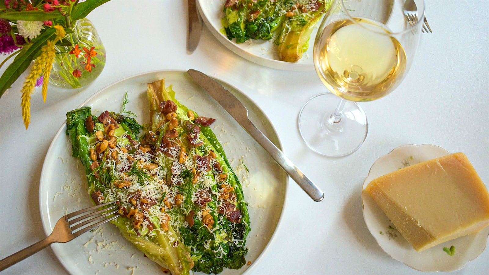 Plates of romaine hearts with yogurt-herb dressing next to a bouquet of flowers, parmesan cheese and a glass of white wine