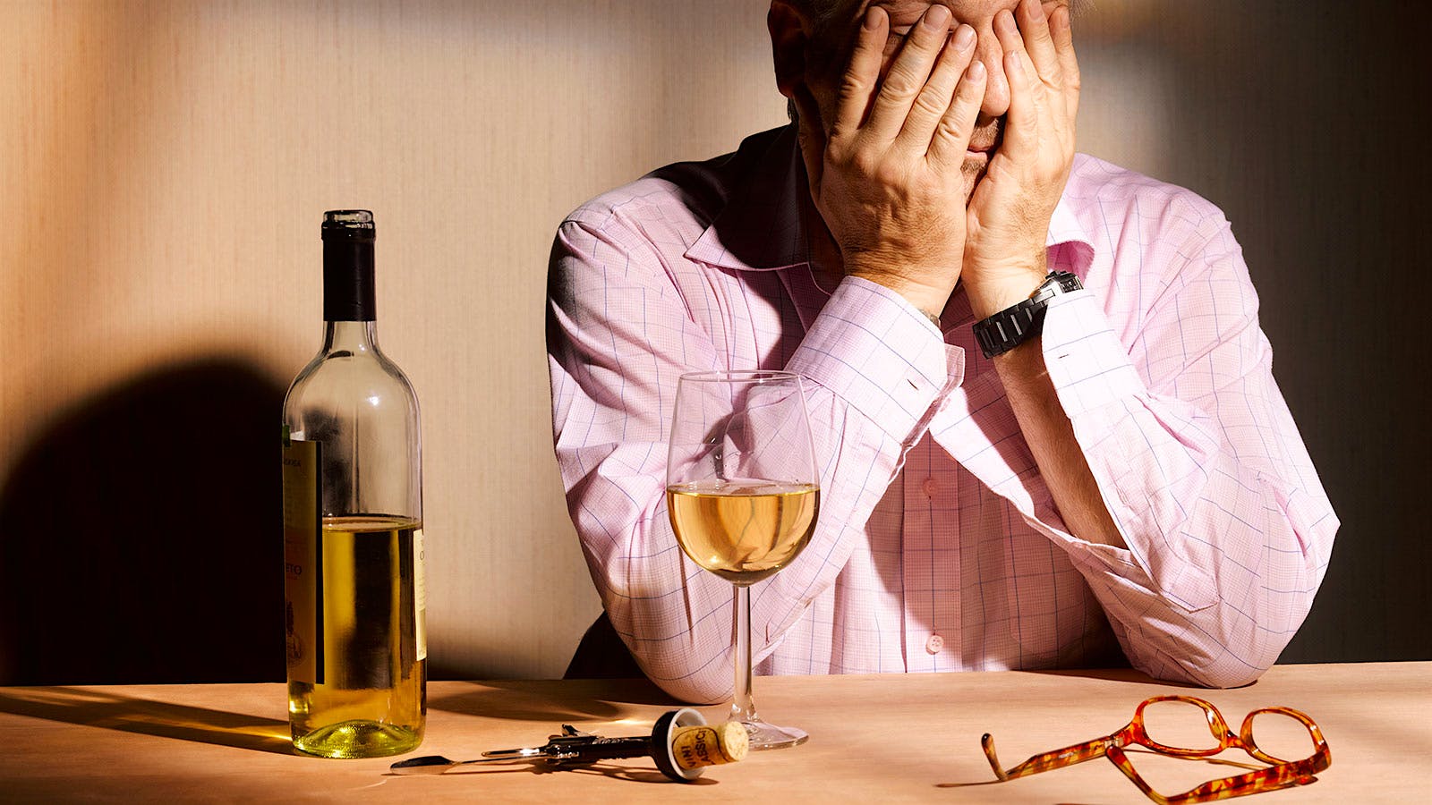 Man holding his hands in his face next to a bottle and glass of white wine