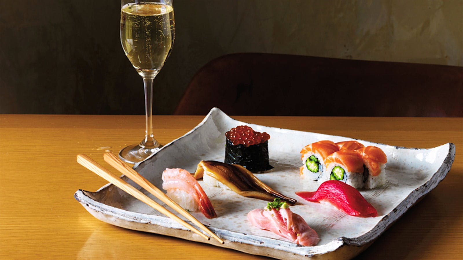 A flute of sparkling wine and a white plate with 6 pieces of sushi and a pair of chopsticks