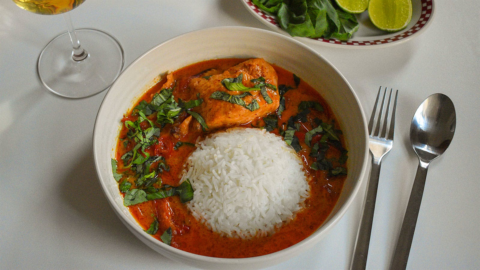 Bowl of Thai red curry salmon and rice next to a glass of white wine and a plate with limes and basil