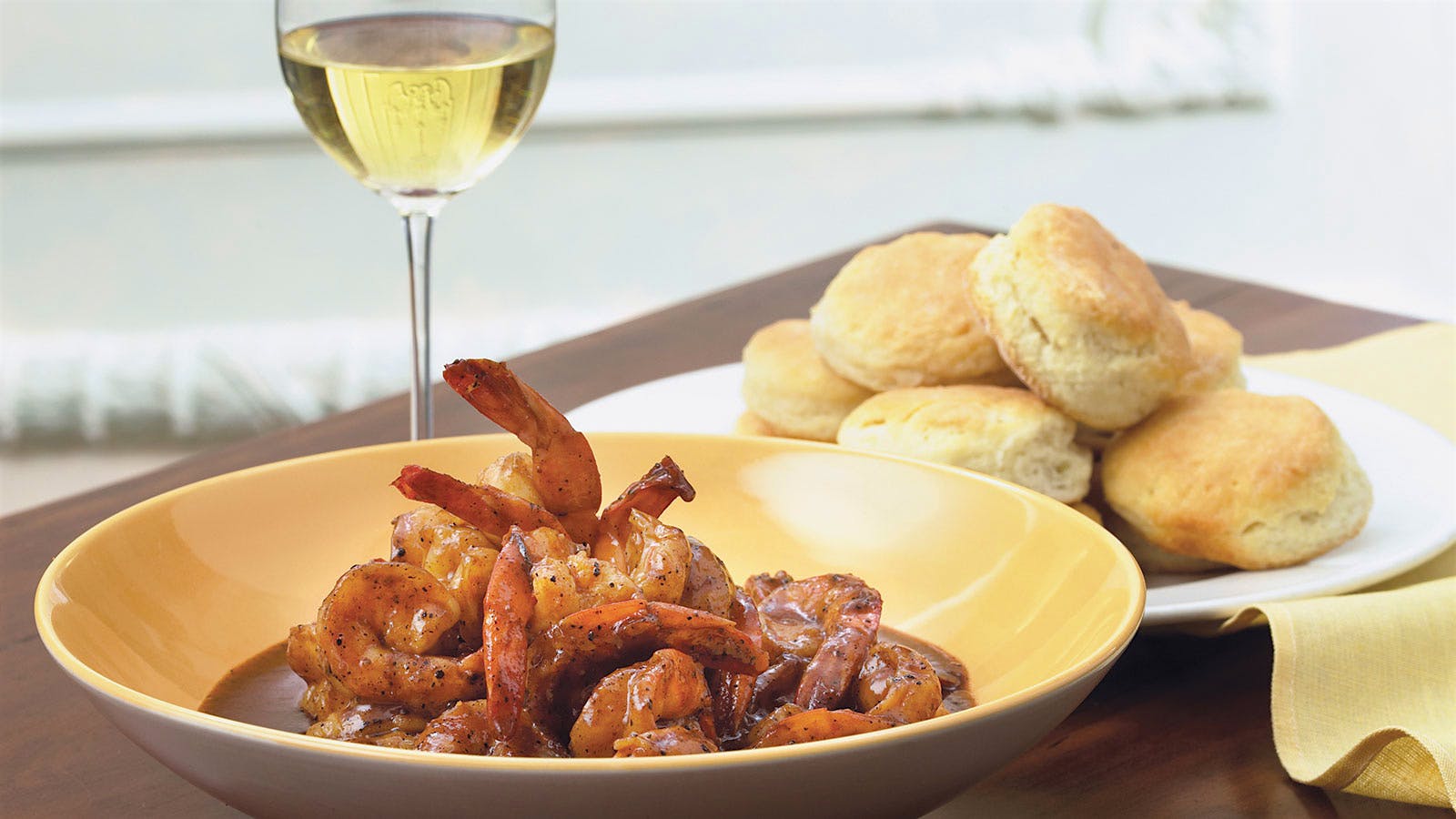 A plate of barbecued shrimp with a plate of biscuits in the background, with a glass of white wine to the left