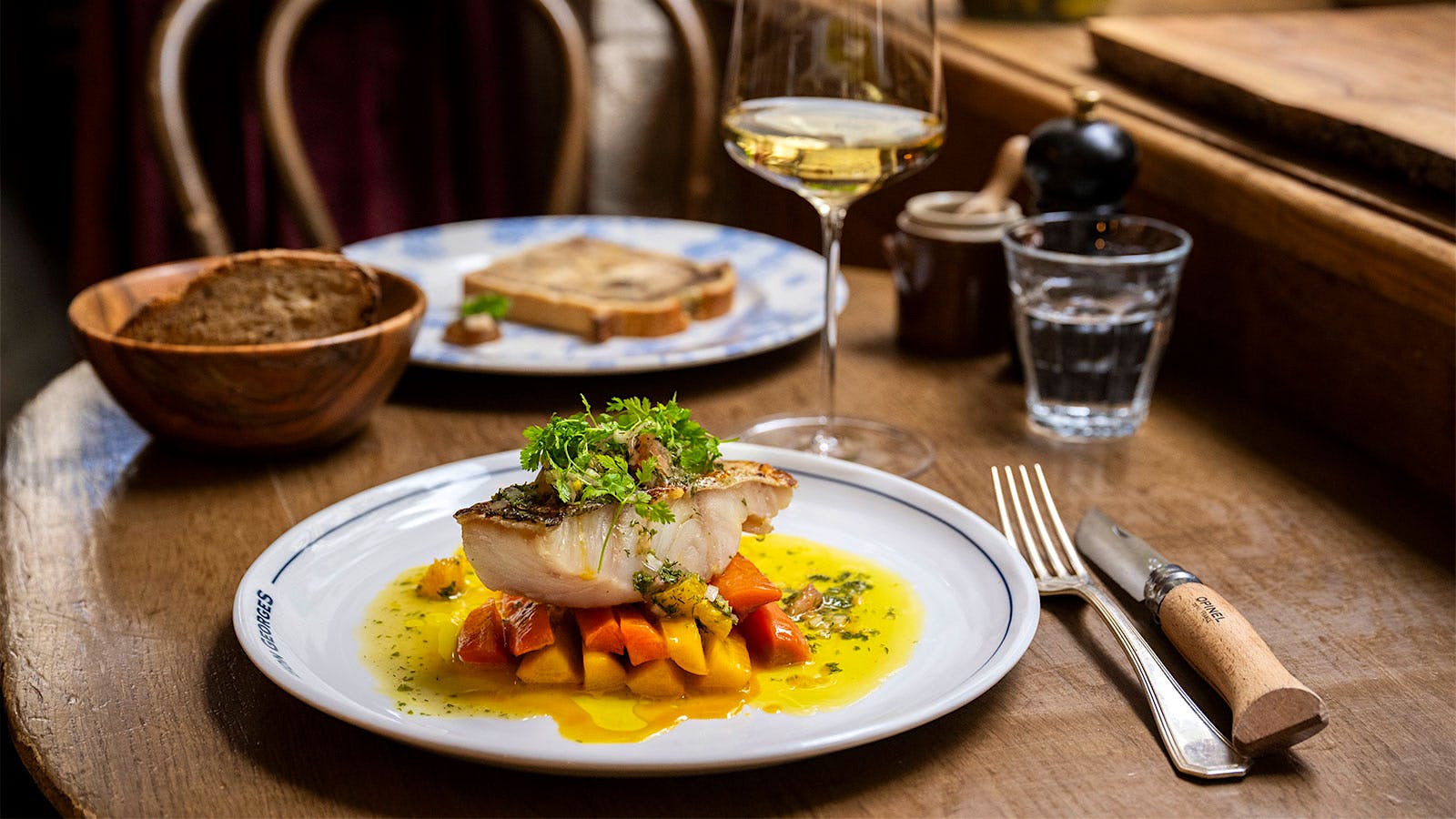  A plate of Le Bon Georges&rsquo; line-caught pollock with glazed carrots and citrus sauce vierge, accompanied by a basket of bread and a glass of white wine