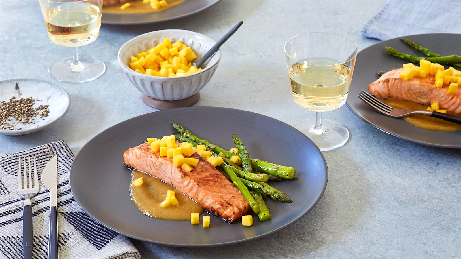A plate of miso-mango salmon topped with quick-pickled mango with a side of roasted asparagus is displayed alongside a glass of white wine