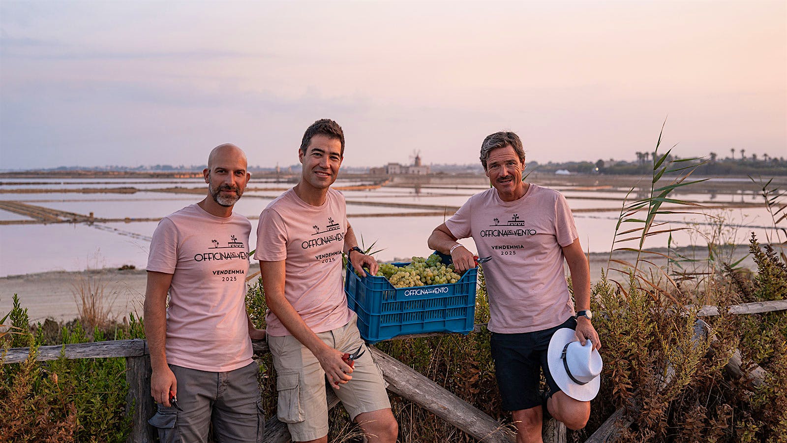  Pietro Russo, Gabriele Gorelli and Andrea Lonardi holding a basket of grapes in front of a landscape of salt flats with a wooden windmill in the background