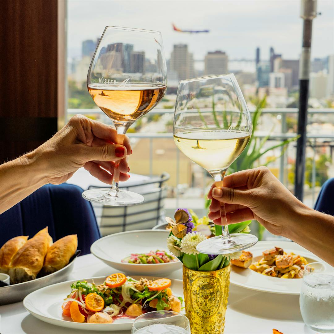 Two diners clink glasses of wine over a table laden with plates of food, overlooking a window with a view of San Diego&rsquo;s harbor, California.