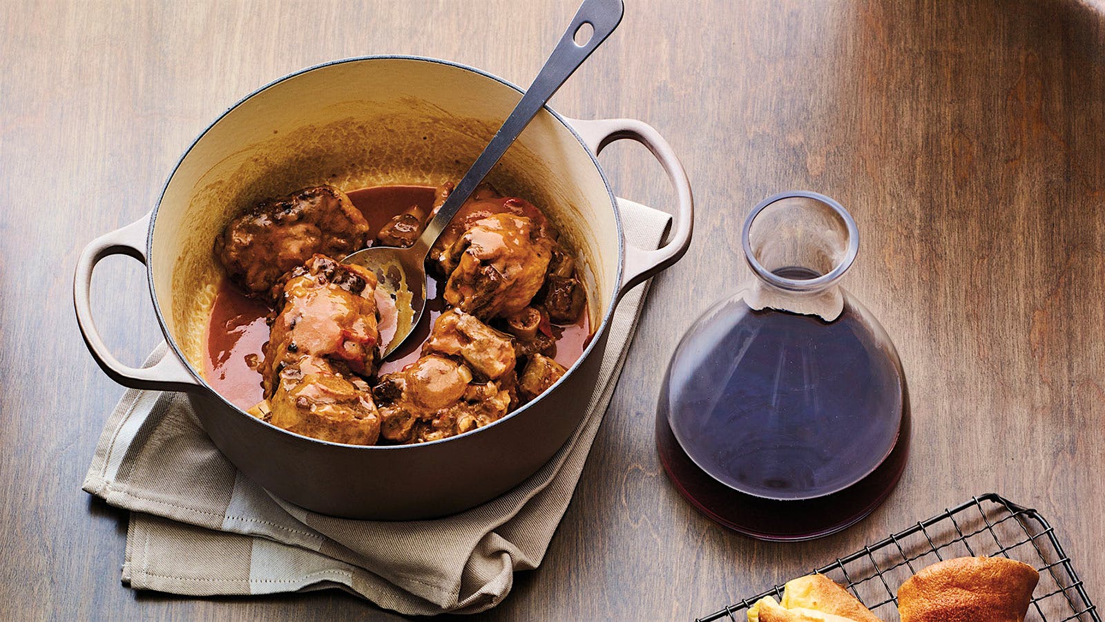 A Dutch oven containing slow-cooked oxtail in a dark pan sauce, accompanied by popovers and a decanter of red wine.