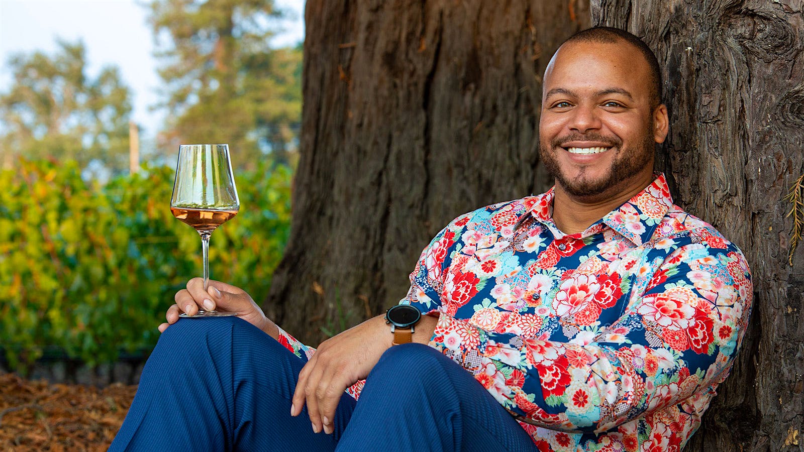 Portrait of Isaiah Fitzgerald-Palacio seated, leaning against a tree, with glass of wine in hand.