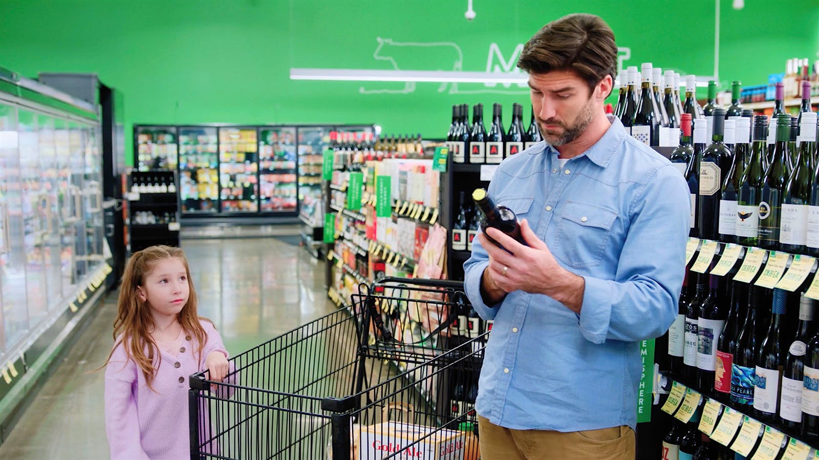 A man inspects a bottle of wine in a supermarket aisle, with a child to his left.