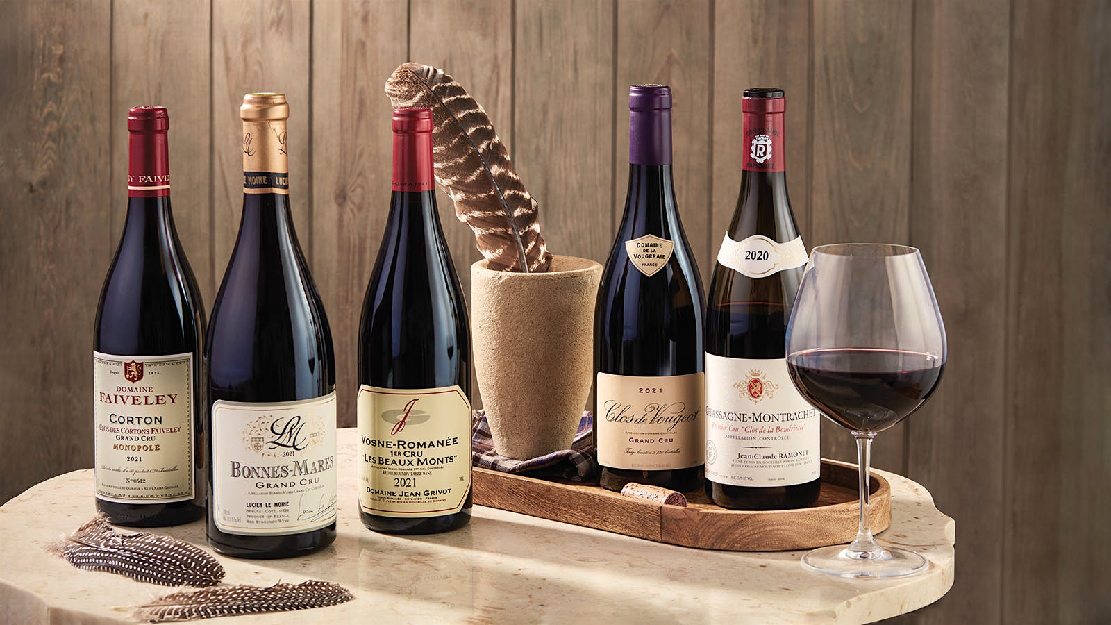 Five bottles of red wines from different Burgundy appellations are displayed side by side on a wooden table next to a half-filled glass and feather in a wooden cup, all sitting in front of a wood-paneled wall.