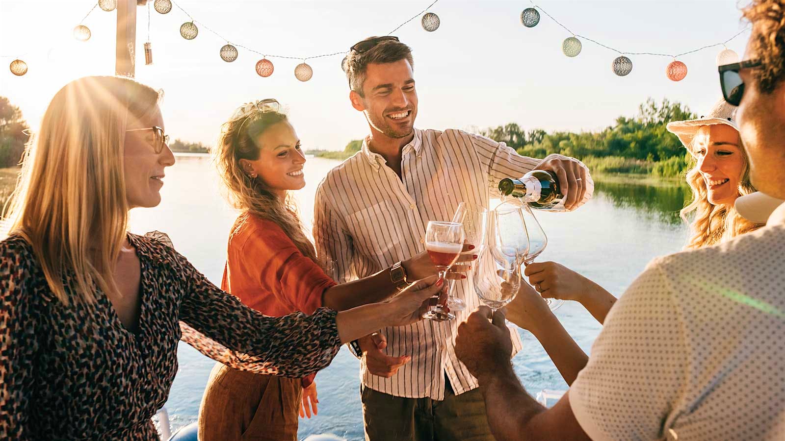 A group of people by a body of water beneath a bunting gather around a bottle of sparkling wine as it is poured into glasses.