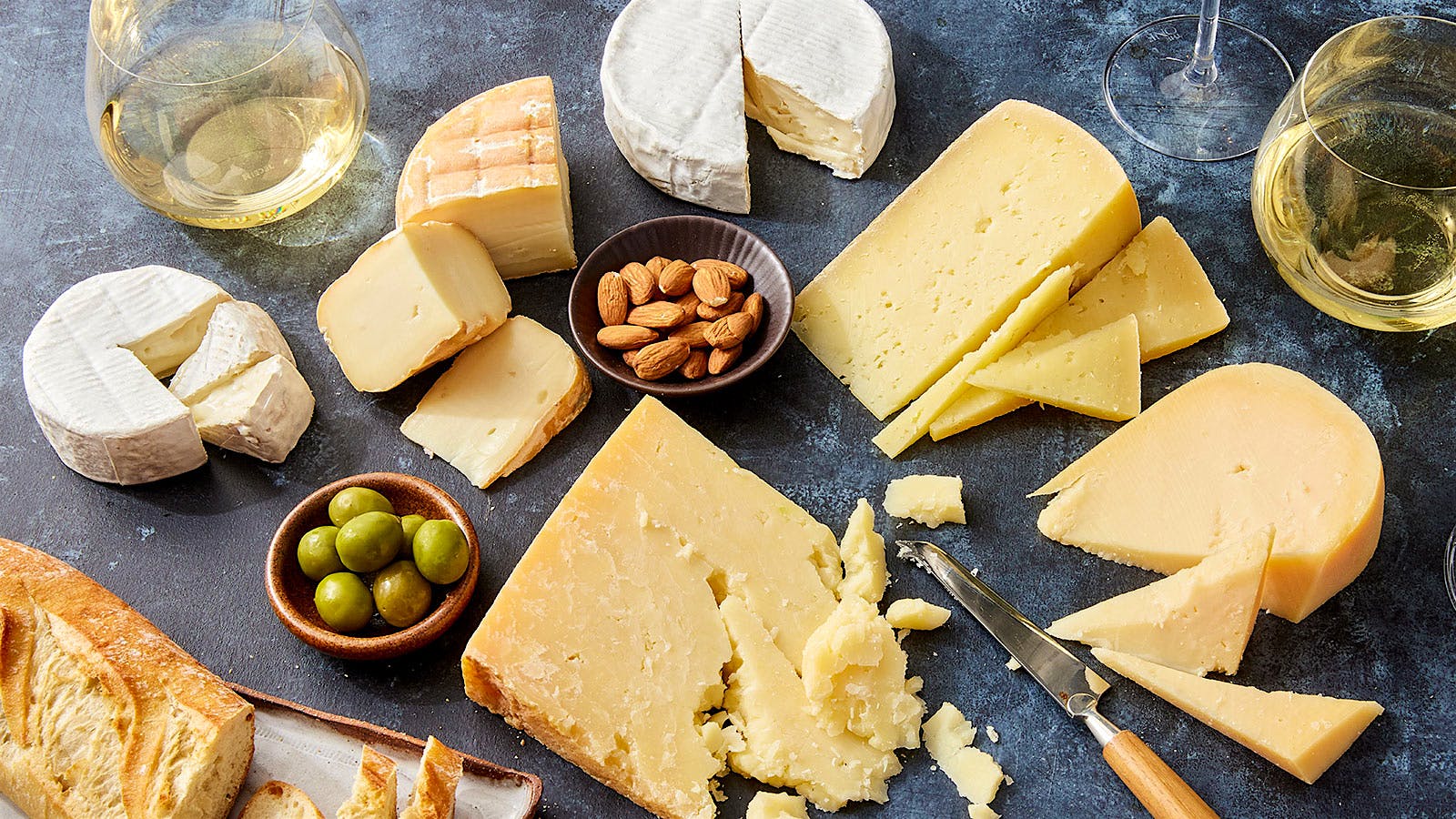 A selection of California cheeses are displayed alongside bowls of olives and nuts, a partially-sliced baguette and two glasses of Chardonnay.